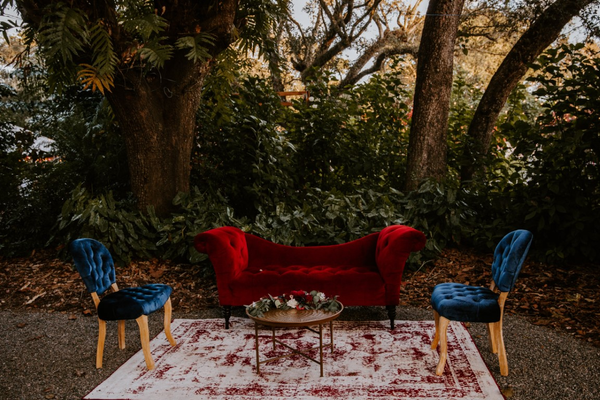 Vintage velvet tufted ruby red assymetrical sofa with wood legs in a lounge set with velvet blue tufted vintage chairs, a red patterned rug, and a bronze gold coffee table. 