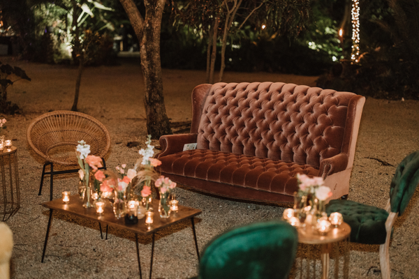 Wood rectangular coffee table with black metal legs centered in a lounge outdoors with flickering candles and florals on top it. A velvet tufted orange dusty rose sofa is behind it. A boho rattan round chair is on one side of the table and on the other si