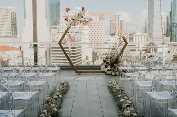 light wood geometric arbor outdoors on tile flooring with a city view of buildings as the background. The arbor is decorated with floral arrangements wrapping around it. In front of it are rows of clear ghost chairs with floral green arrangements. 