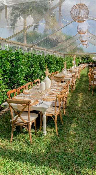 Wooden white rectangular farmhouse farm table with white turned wooden legs with wood crossback chairs on grass with clear tent overhead. Circular boho decor hanging.