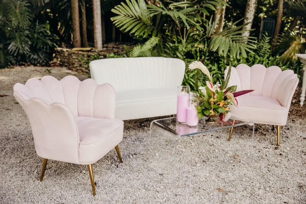 A vintage-inspired outdoor seating arrangement with a white sofa and two blush pink tufted armchairs, accented by a glass coffee table and tropical foliage in the background.