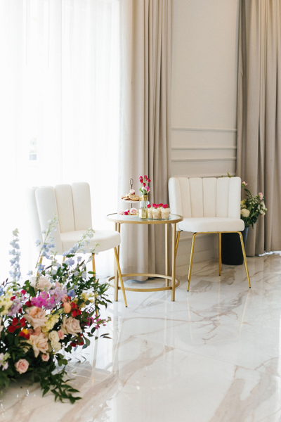 Modern velvet ivory beige chairs with gold legs on each side of a gold round table with a floral arrangement in front of it. 