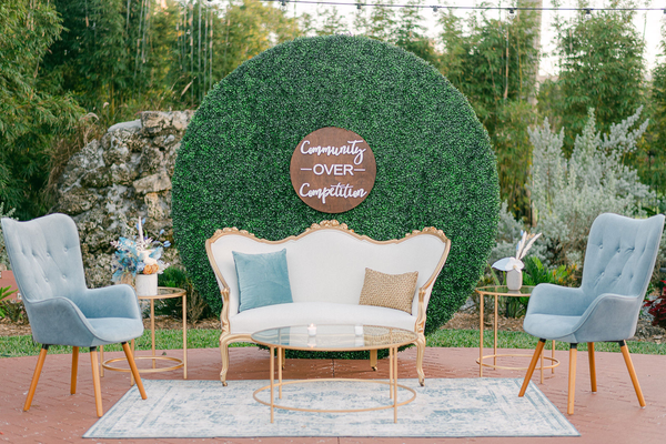 Vintage gold round side table in a lounge set including a vintage and gold loveseat, tufted blue chairs, a round gold coffee table and a blue rug. In the background there is a round grass backdrop with a wood sign outdoors. 