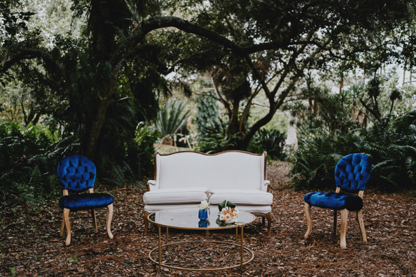 Modern vintage linen ivory beige loveseat with wood accents in a lounge set with blue vintage tufted chairs and a round gold coffee table outdoors with trees in the background.