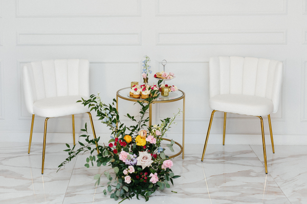 Modern velvet ivory beige chairs with gold legs on each side of a gold round table with a floral arrangement in front of it. 