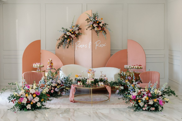 An elegant event space is defined by a central white sofa flanked by peach-colored circular backdrops with floral accents and personalized text, complemented by vibrant floor floral arrangements.