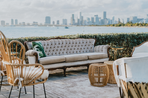 boho rattan round geometric side table in a lounge set including a large tufted vintage sofa, boho rattan chairs overlooking the ocean with a city view and a bush fence nearby.