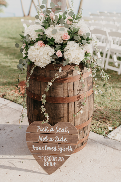 Rustic farmhouse dark wood round barrel with florals on top of it and a heart shaped sign at the base on the floor with writing near white chairs. 