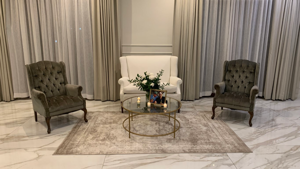 ivory beige loveseat with a gold round coffee table with vintage velvet green arm chairs with wood legs on either side of the coffee table on a beige ivory rug indoors. 