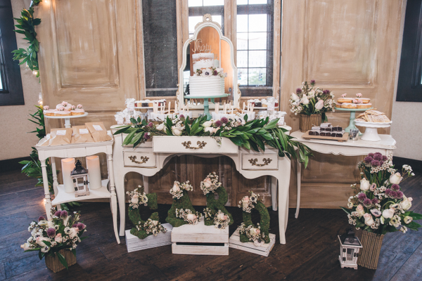 Vintage ivory beige rectangular accent table indoors on wood flooring with a wood door panel as a backdrop behind it. Green plants with florals are decorating the table with a tiered cake. Next to it are other vintage tables with trays with sweets on each