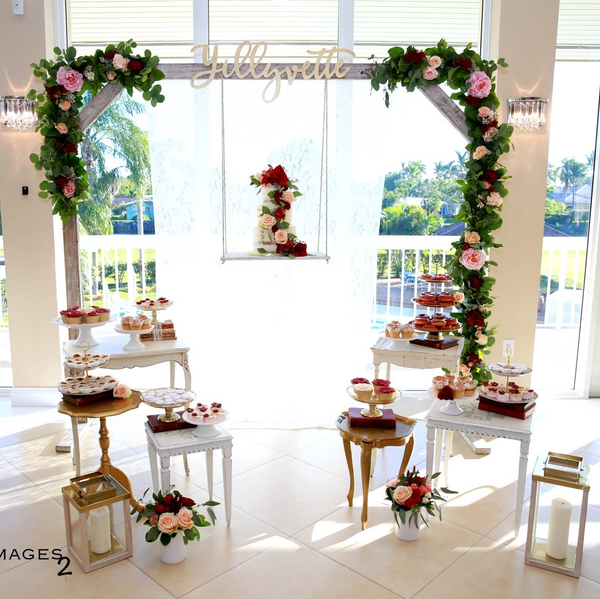 Farmhouse whitewash wood square arbor indoors with green and pink floral garland with a swing hanging from it where a white and red tiered cake sits. Small accent tables surround it with trays of sweets on all of them. 