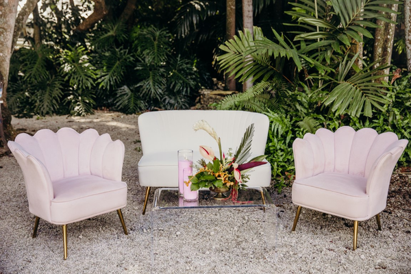 Modern linen ivory beige loveseat with gold legs with pink floral shaped chairs with gold legs on each side. An acrylic clear coffee table in the center with a floral arrangement outdoors. 