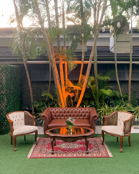 Vintage brown wood oval coffee table centering a lounge outdoors over a patterned red rug and with a small floral arrangement on it. A vintage tufted leather brown sofa behind it and two tufted wood and velvet arm chairs on each side of the table. Behind 