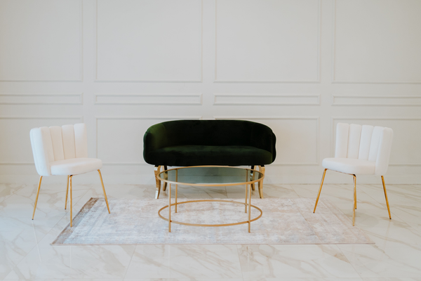 A minimalist setting with a modern black velvet bench centered between two white chairs with gold accents, all poised around a sleek, low-profile glass coffee table.
