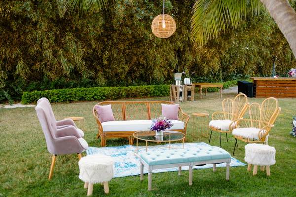 lounge set with rattan boho sofa with a gold round coffee table with rattan boho floral shaped chairs on one side of the coffee table on a blue patterned rug and pink tufted chairs on the other. A vintage green bench and bohoottoman on the edge of the rug