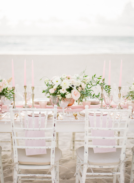 White distressed chiavari chairs with white  legs tucked into a white wood farm table decorated with florals and candles on a sandy beach overlooking the ocean. 