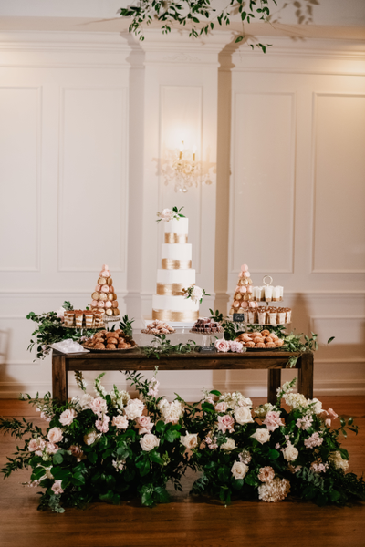 Dark wooden rectangular brown farm house farm table with dark brown wooden legs with floral decor at the base of it and trays with desserts and a tiered cake on top of it. 