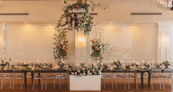 Dark wooden rectangular brown farm house farm table with dark brown wooden turned legs with clear ghost chairs tucked into it with tableware and floral on it. A large hanging floral arrangement overhead hanging on the roof.  A white blocked table in the c