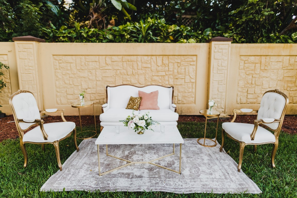 ivory beige loveseat with a gold round coffee table with a tufted arm chairs on either side of the coffee table on a beige pattern rug outdoors.