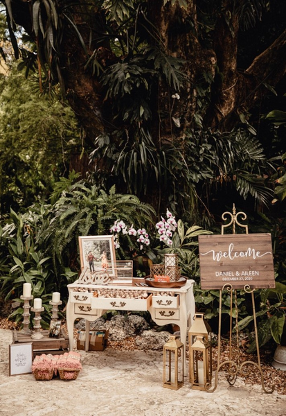 Vintage gold easel sign holder with a large sign with writing next to a dresser table with a frame and floral vases on it and small decor and candles on the corner base of it on the floor outdoors. Large trees are in the background.