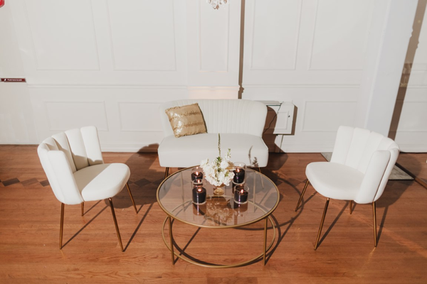 A modern gathering space with three white mid-century chairs arranged around a round table with a geometric gold base, featuring a centerpiece of flowers and candles.