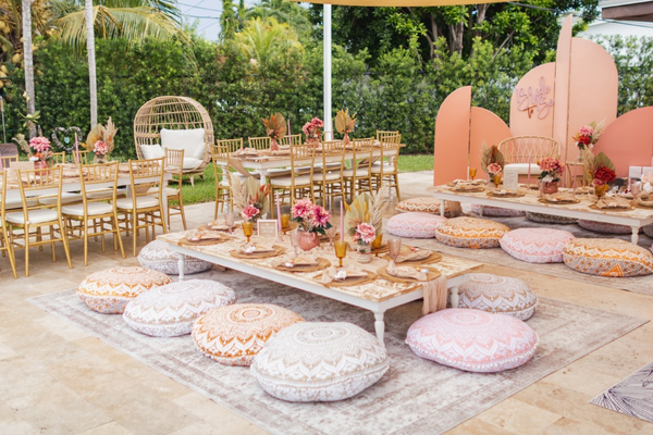 Wooden white distressed rectangular farmhouse boho picnic table with white wooden turned legs with boho floor patterned pillows with boho decor and tableware on top of it. A large asymmetrical pink background backdrop with the lounge in the corner. Dining