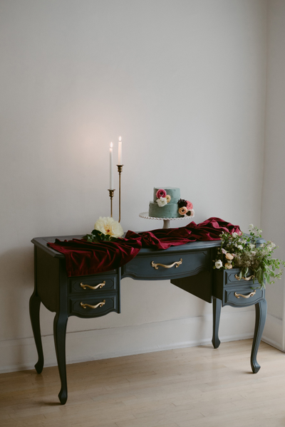 Vintage indigo blue rectangular dresser accent table with gold accents with red draping over the top. Two long lit candles on candle holders are on top of the table next to araised cake stand with a blue cake that has floral decor. 