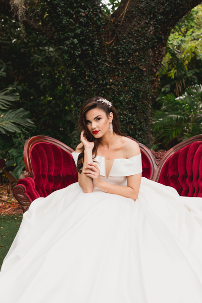 Vintage velvet tufted burgundy red loveseat with dark brown wood accents with a woman in a dress sitting on it in front of a large tree outdoors. 