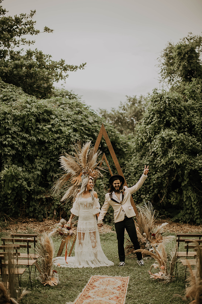 Boho wood triangle arbor with a bride and groom standing in front of it while the groom raises his hand and the bride smiles. There are chairs surrounding them and a rug infront of them. Boho decor decorates the arbor.