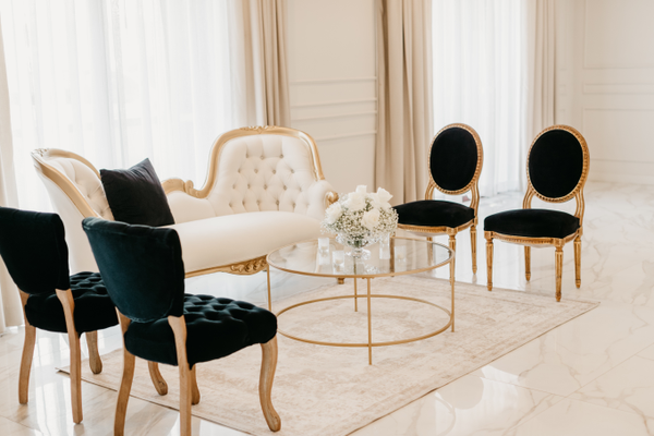 Vintage gold and ivory beige sofa with a modern gold and glass coffee table with a black vintage chairs on each side of the coffee table over a beige rug.