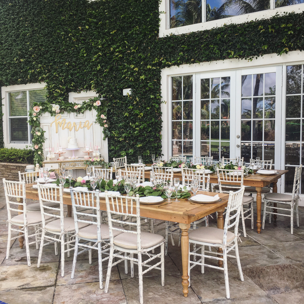 White distressed chiavari chairs with white  legs tucked into a wood farm table with a green grass garland draped over it with tableware and candles decorating it. A grass wall and window doors are behind it. 