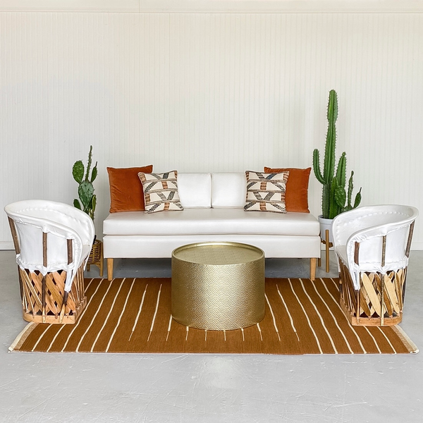 ivory beige sofa with a gold round coffee table with rattan boho and white chairs on either side of the coffee table on a rust colored pattern rug with a bench on the edge of it. 