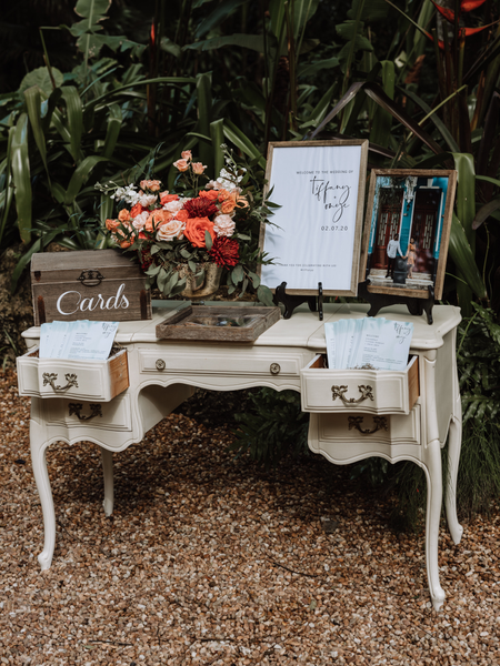 Vintage ivory beige rectangular dresser accent table outdoors with pink and red flowers on top and a wood box and frames surrounding it. The drawers of the table are open with rectangular papers with writing coming out f them. 