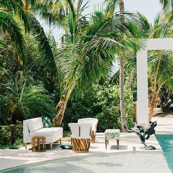 Vintage bronze gold round side table next to a lounge set that includes white rattan chairs, white and wood sofa, and a green palm and white bench with wood legs near a pool overlooking trees.