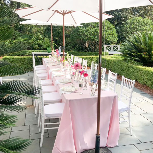 White Chiavari chairs under umbrellas with pink linens on patio surrounded by boxwood for bridal shower.