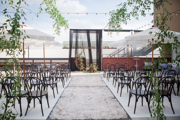 Rooftop wedding with hanging smilax over black bentwood boho chairs with rug as aisle runner.