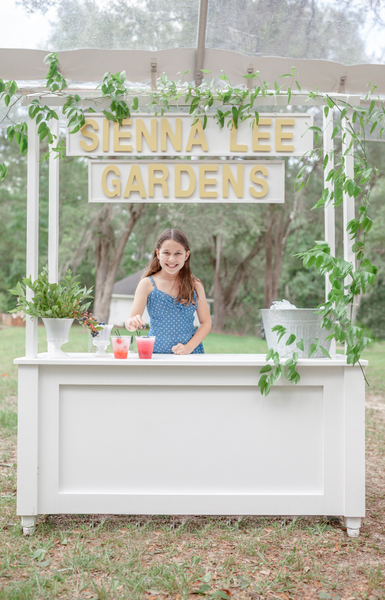 White lemonade stand with smilax and little girl server at Sienna Lee Gardens.