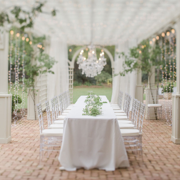 Long white table with acrylic Chiavari chairs under pergola with chandelier at The Space at Feather Oaks.