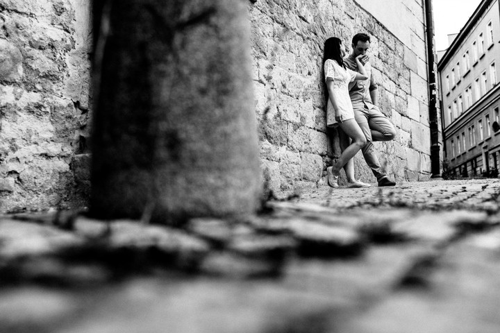 Junges Paar lehnt entspannt an einer historischen Sandsteinmauer in einer kopfsteingepflasterten Gasse, hält Händchen und lächelt sich zärtlich an.