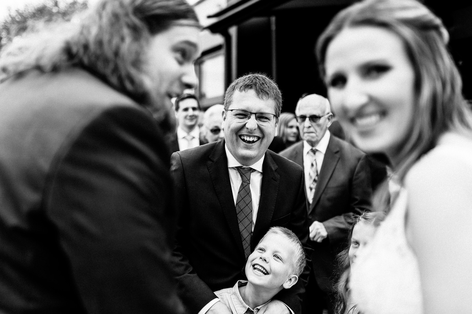 Braut und Bräutigam lachen mit ihren Gästen, während ein kleiner Junge freudig im Vordergrund steht.