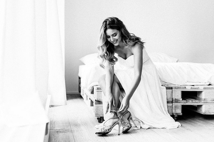 Braut sitzt auf einer hellen, modernen Bettenlandschaft und zieht sich lächelnd ihre Hochzeitsschuhe an.