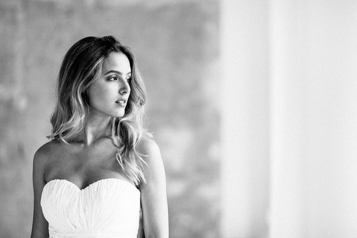 Braut in elegantem, schulterfreiem Brautkleid blickt verträumt in die Ferne, mit weichem Licht und heller, minimalistischer Kulisse.