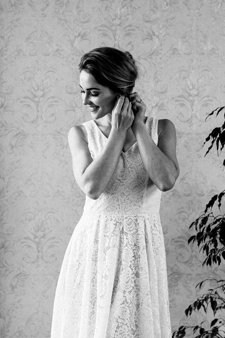 Braut in elegantem Spitzenkleid, die lächelnd ihre Ohrringe anlegt, vor einer dezenten Tapete mit Pflanzen im Hintergrund.