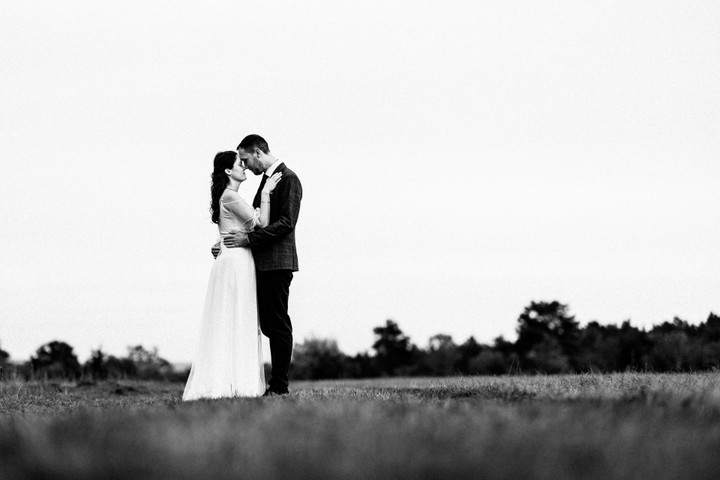 Braut und Bräutigam stehen inmitten einer weiten Wiese im sanften Abendlicht und tauschen einen zarten Kuss, sie im fließenden Kleid, er im eleganten Anzug.