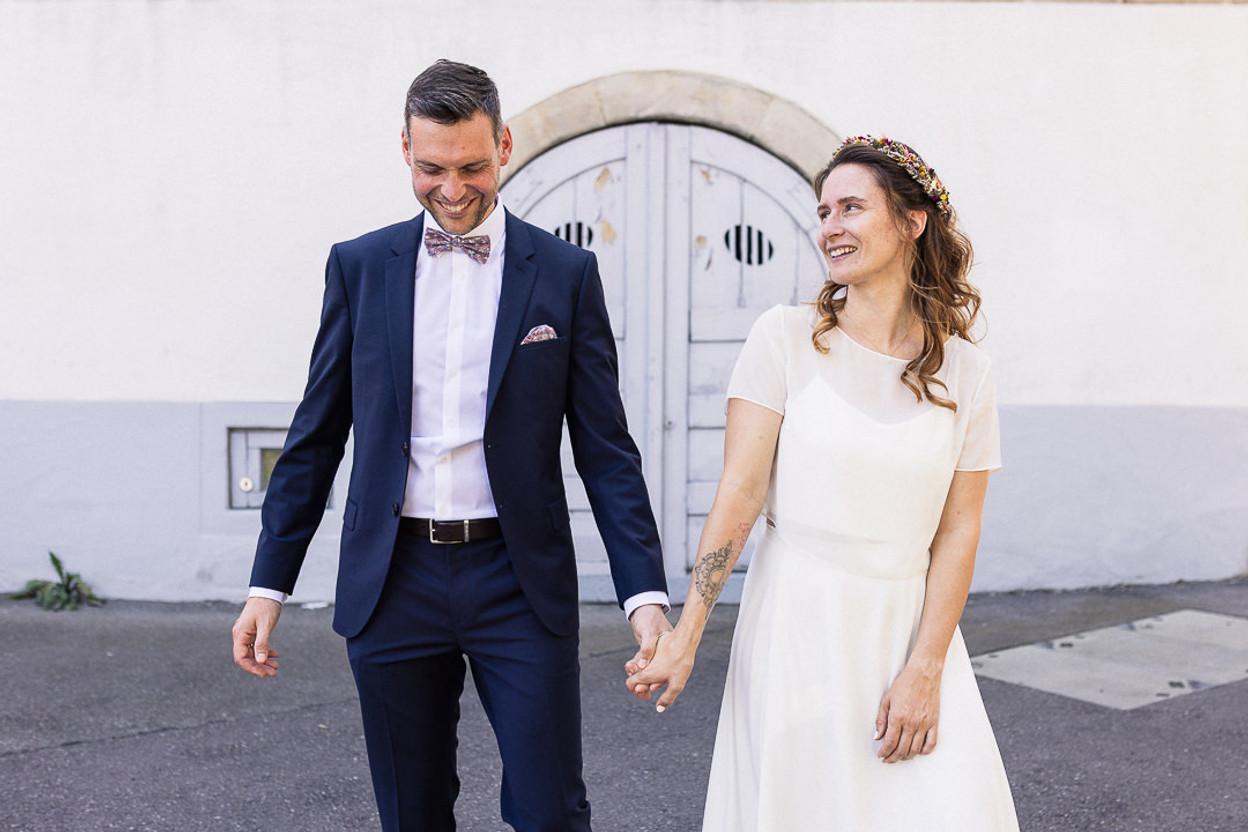 Frisch verheiratetes Paar nach dem Standesamt in Böblingen – der Bräutigam im dunkelblauen Anzug und die Braut im schlichten weißen Kleid mit Blumenkranz lachen einander an und halten sich an den Händen.