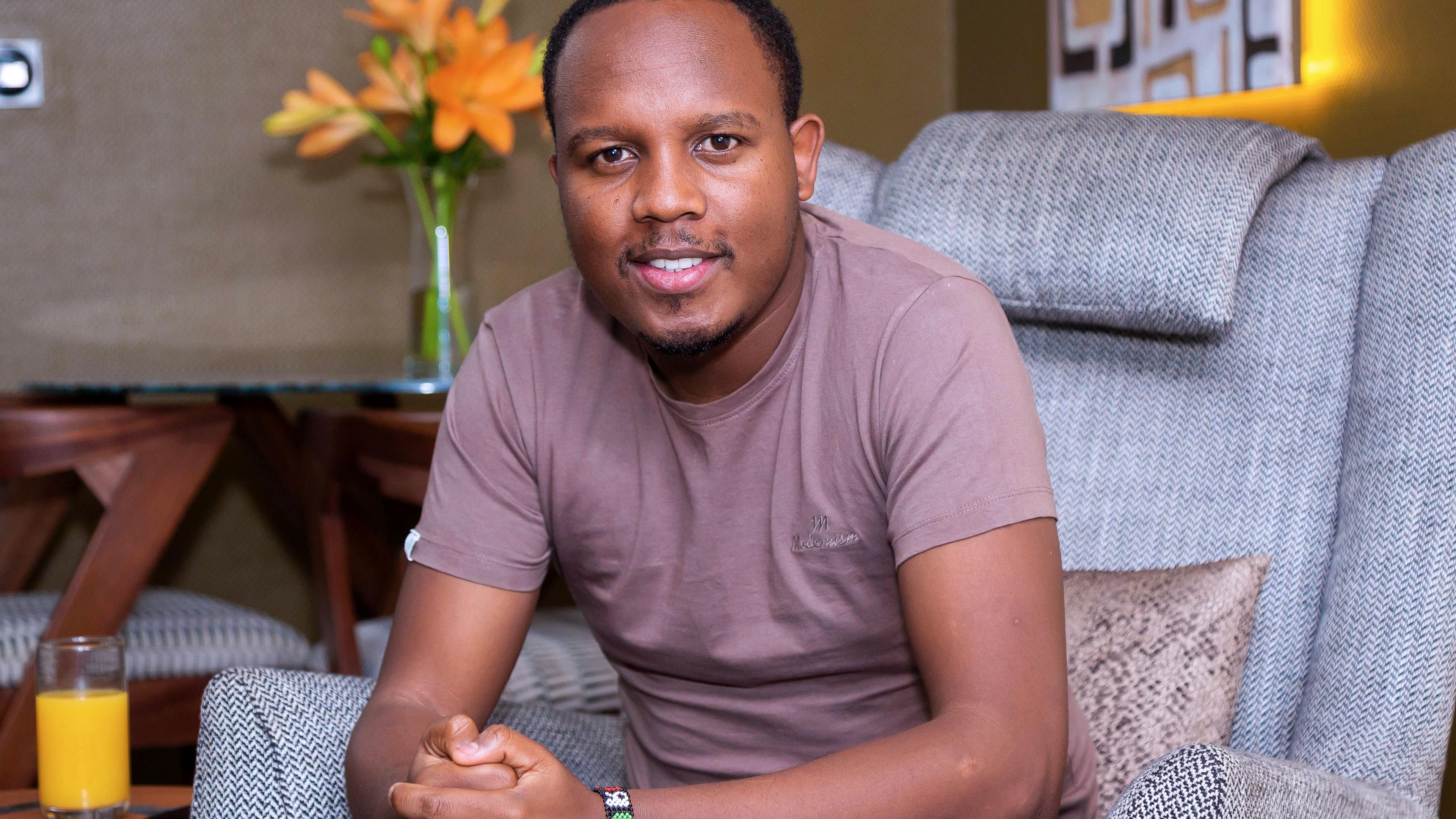 A close up image of a man sitting on a sofa facing the camera, sporting an armband and hands held together