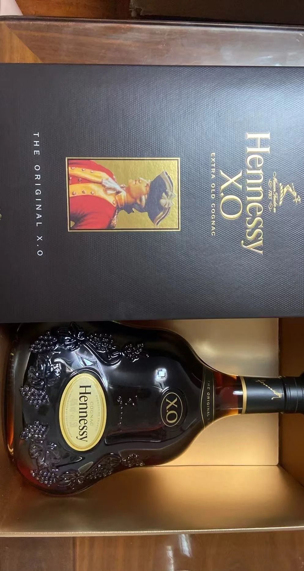 軒尼詩 XO 酒瓶橫放在盒裡,盒蓋在後方。瓶頸是黑色頸帶,瓶標寫著 Hennessy 與 XO。