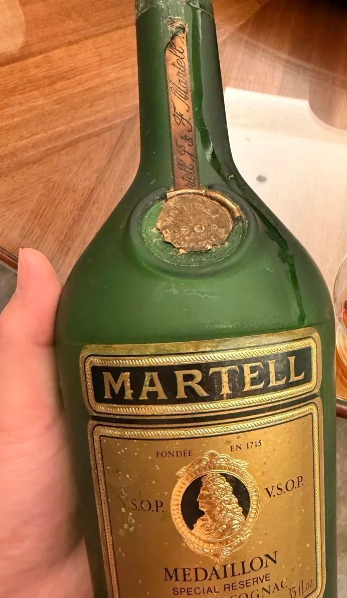 一隻手握著綠色酒瓶。酒標寫著 MARTELL、V.S.O.P.、MEDAILLON 與 SPECIAL RESERVE。瓶肩有一條磨損的紙條和一枚圓形印章。瓶蓋不在畫面內。