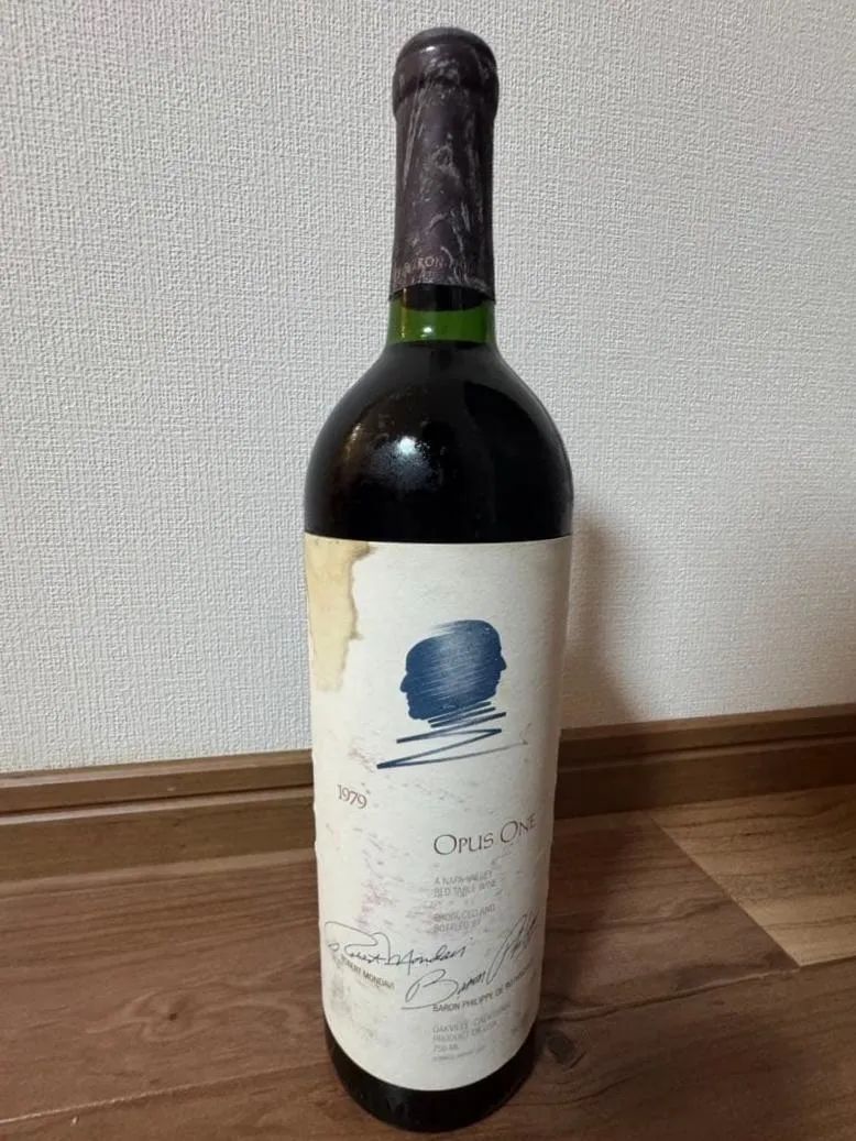 一支直立酒瓶,酒標可見 1979、OPUS ONE、藍色側面人像和兩處簽名,酒標左側污漬並撕裂,瓶帽深色且風化,液位在瓶頸