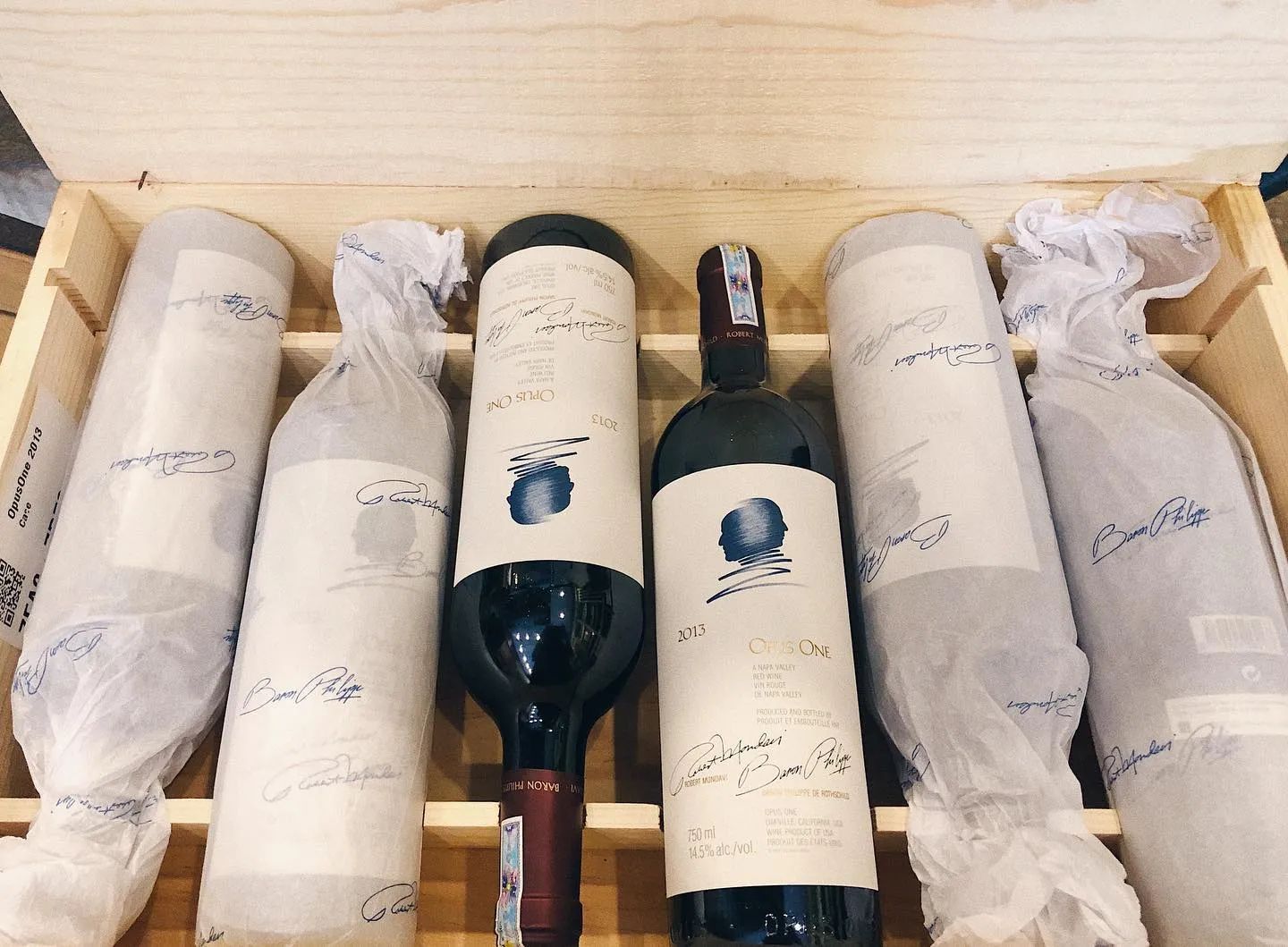 打開的木箱裡放著酒瓶,兩支未包紙的瓶子酒標可見 OPUS ONE 和 2013,其餘瓶子包著印花薄紙